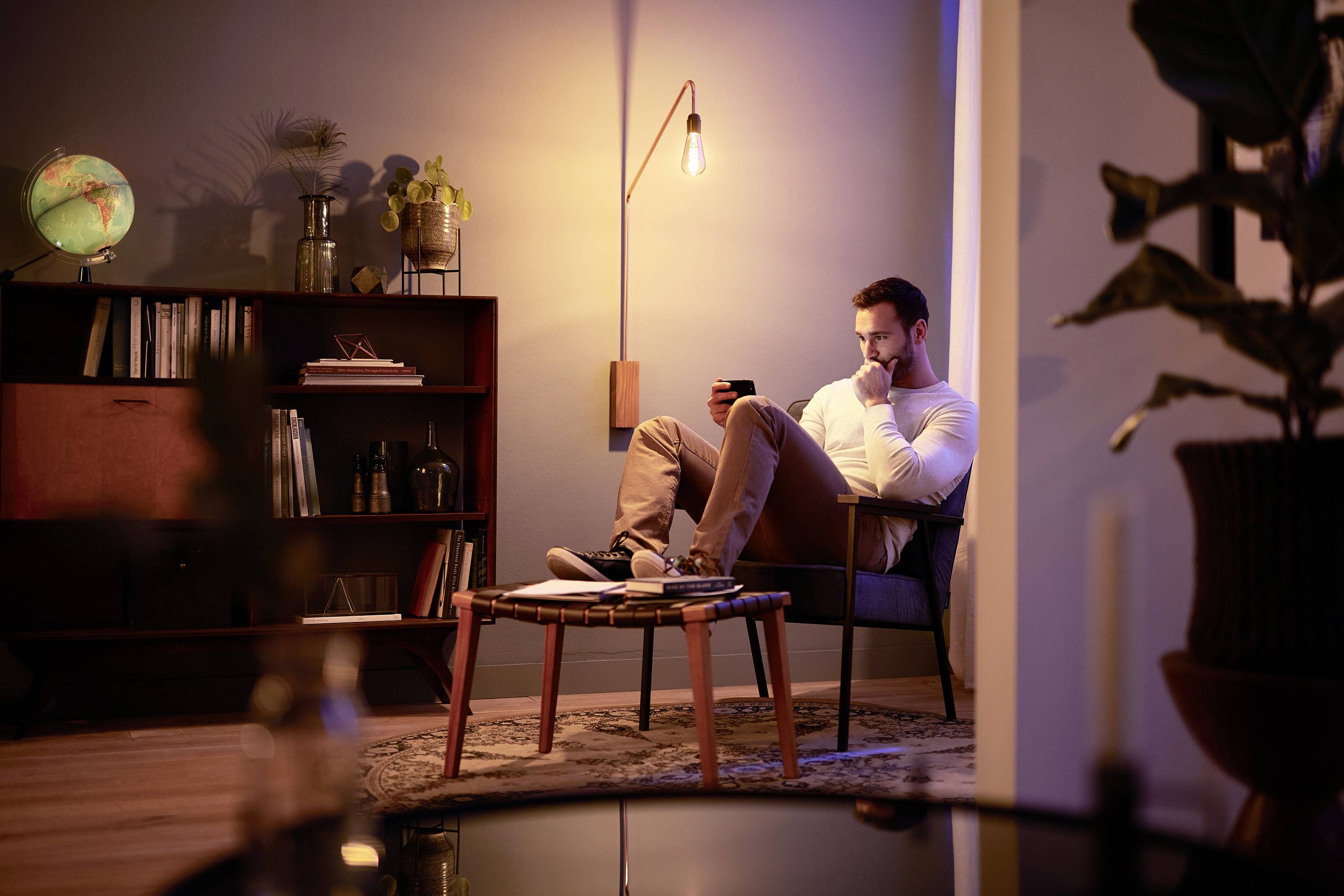 Ein Mann sitzt in einem gemütlichen Raum und liest, umgeben von Bücherregalen, Pflanzen und warmer Beleuchtung.