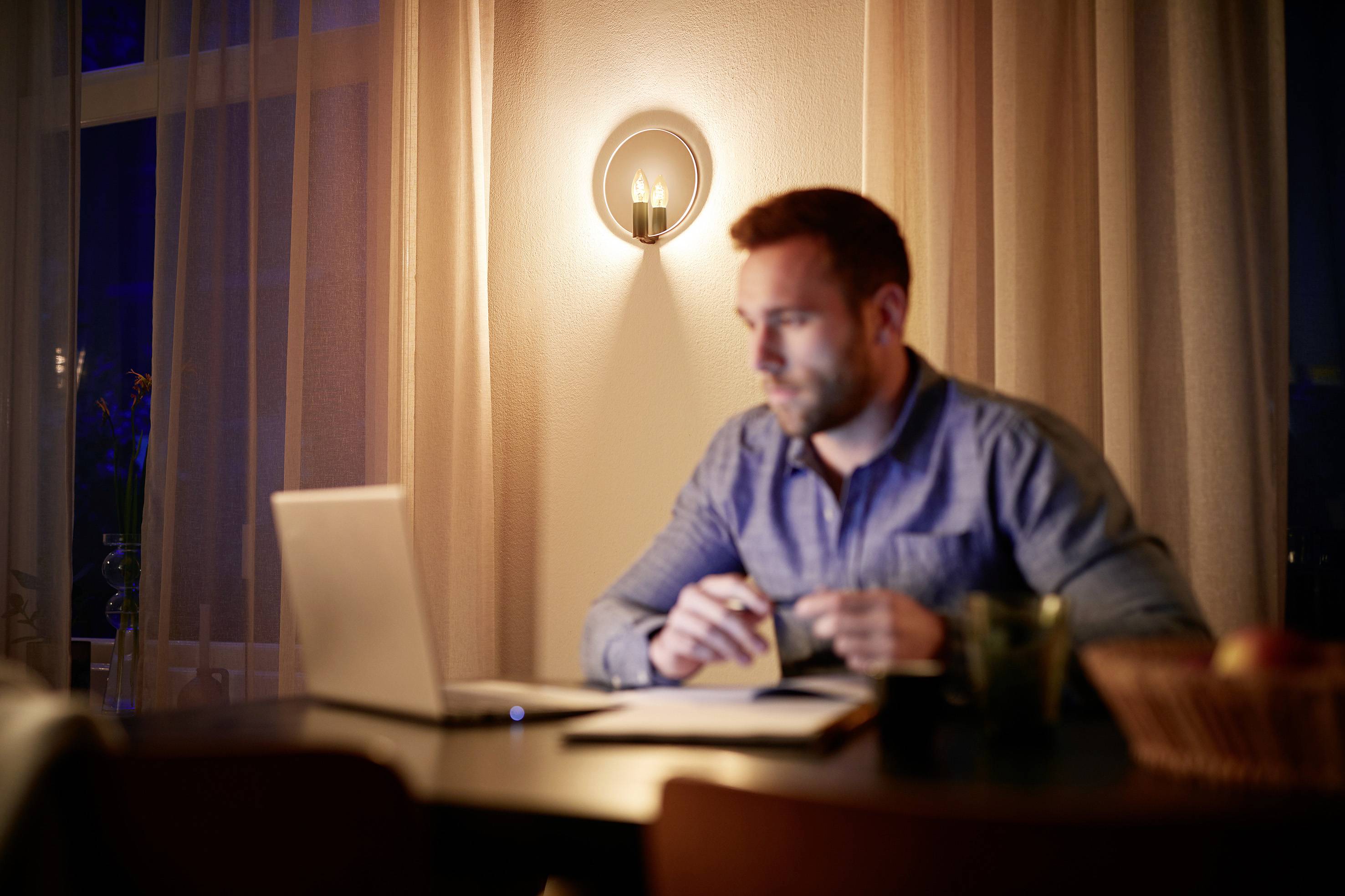 Ein Mann sitzt in einem abgedunkelten Raum an einem Tisch und arbeitet konzentriert an einem Laptop. Im Hintergrund leuchtet eine Lampe an der Wand.