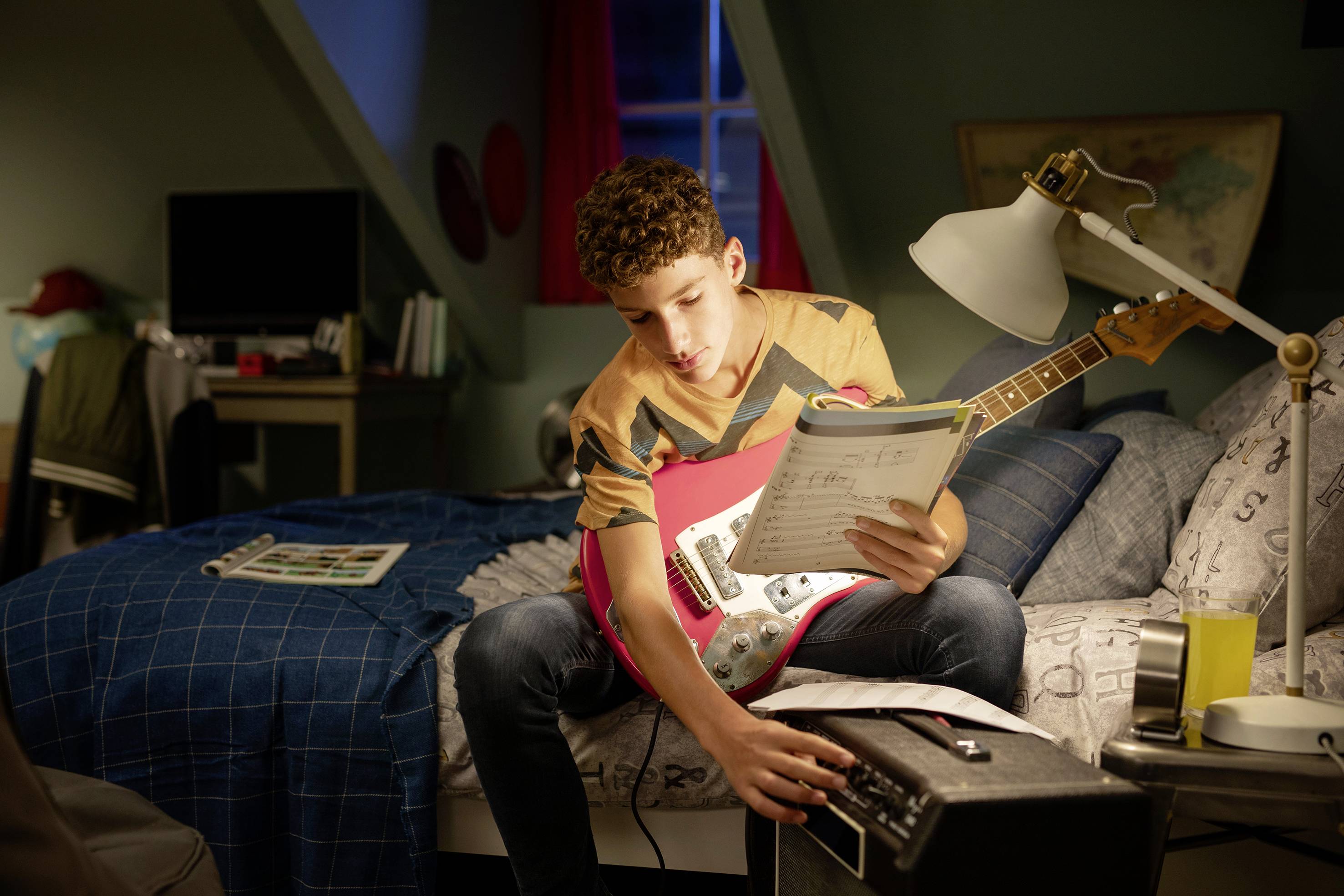 Junge sitzt auf einem Bett, hält eine E-Gitarre und Notenblätter, während er einen Verstärker einstellt. Schlafzimmer mit Schreibtisch im Hintergrund.