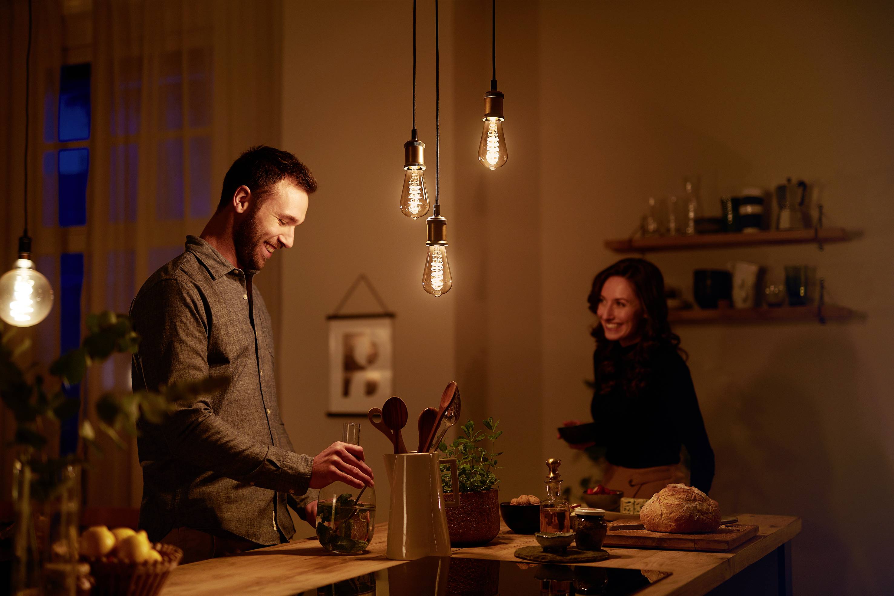 Ein Mann kocht in einer gemütlich beleuchteten Küche, während eine Frau im Hintergrund lächelnd zusieht.