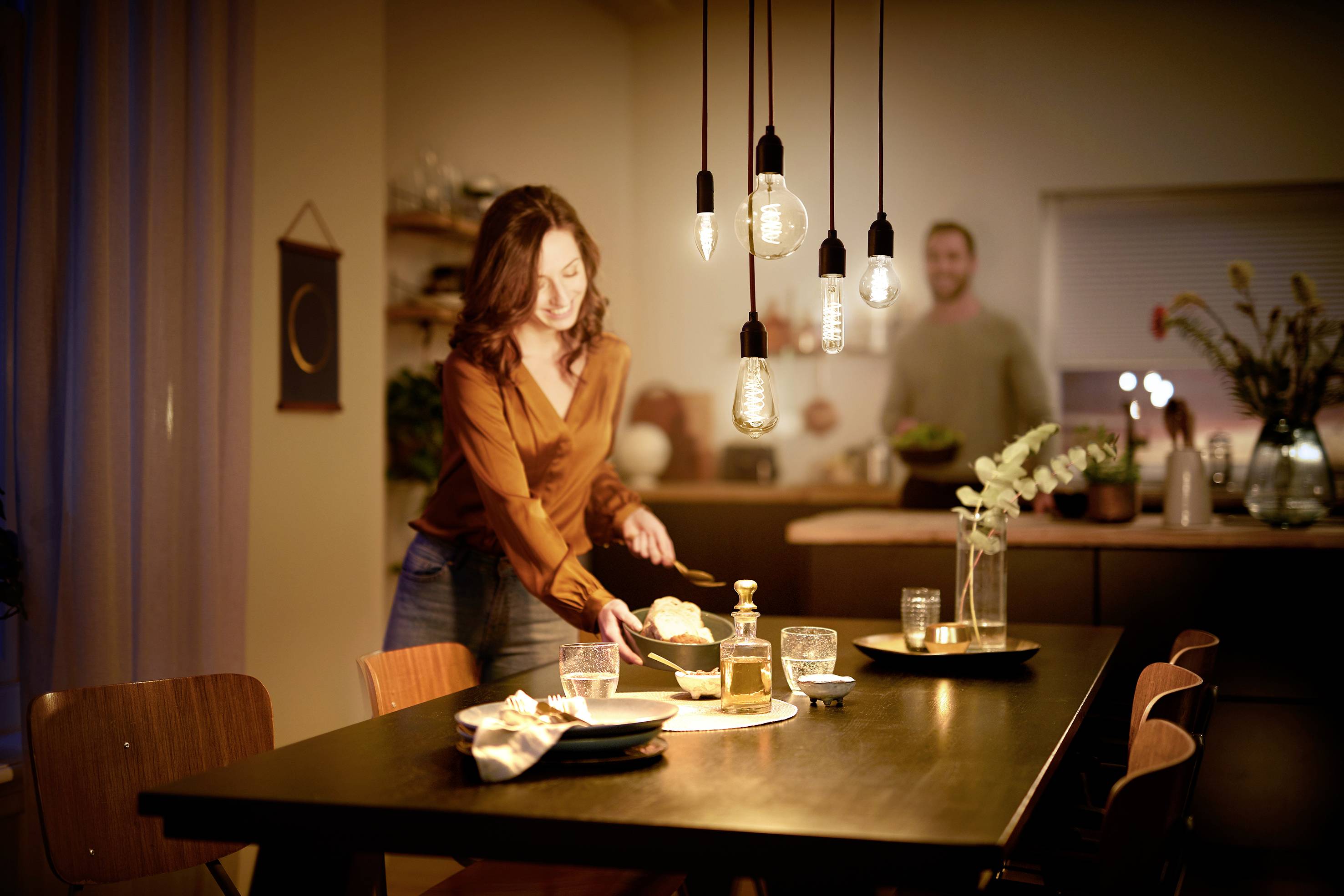 Eine Frau deckt einen Tisch in einem gemütlich beleuchteten Esszimmer. Im Hintergrund steht eine weitere Person in einer Küche.
