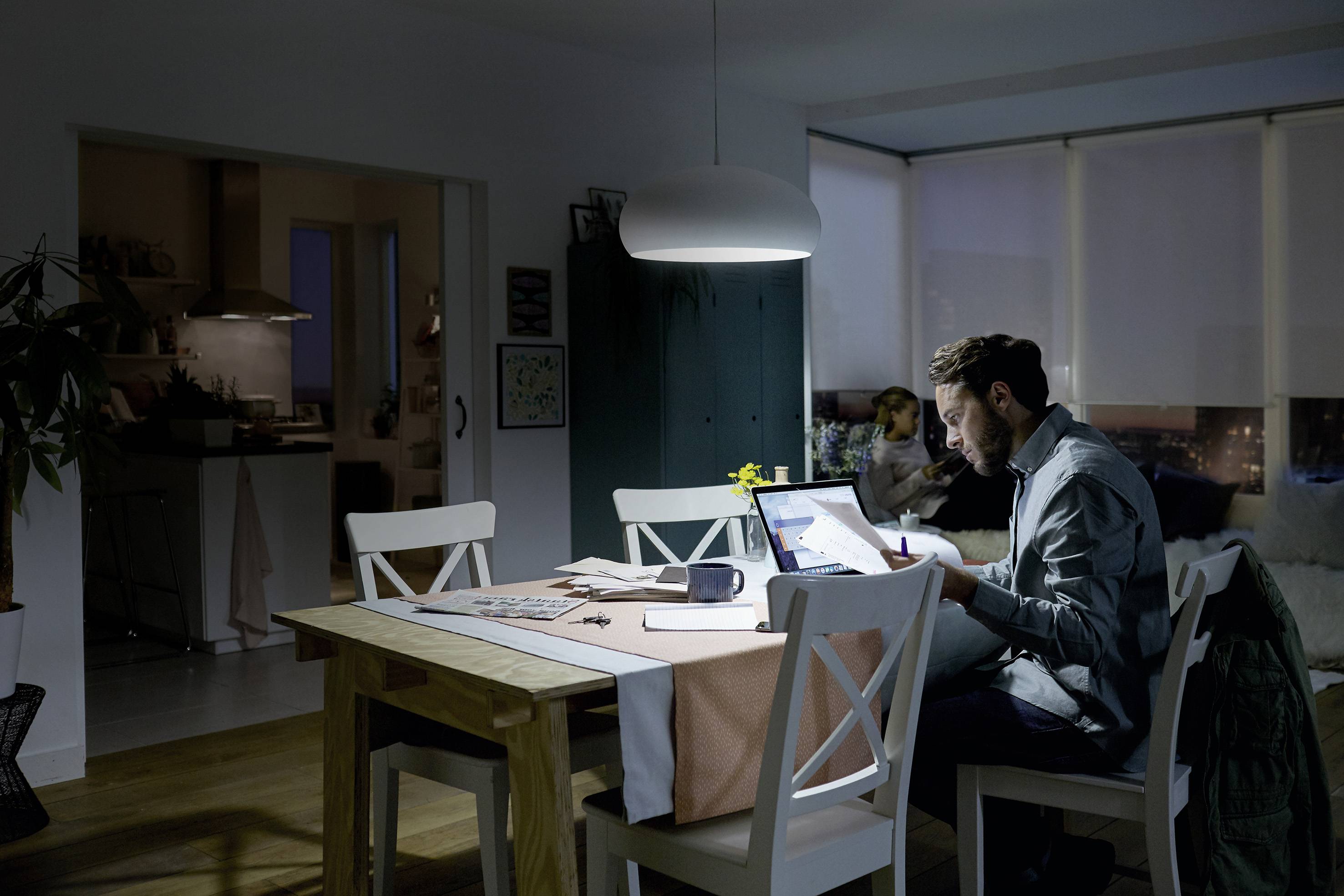 Ein Mann arbeitet spätabends am Laptop in einem dunklen Raum. Ein Kind sitzt im Hintergrund auf dem Sofa.