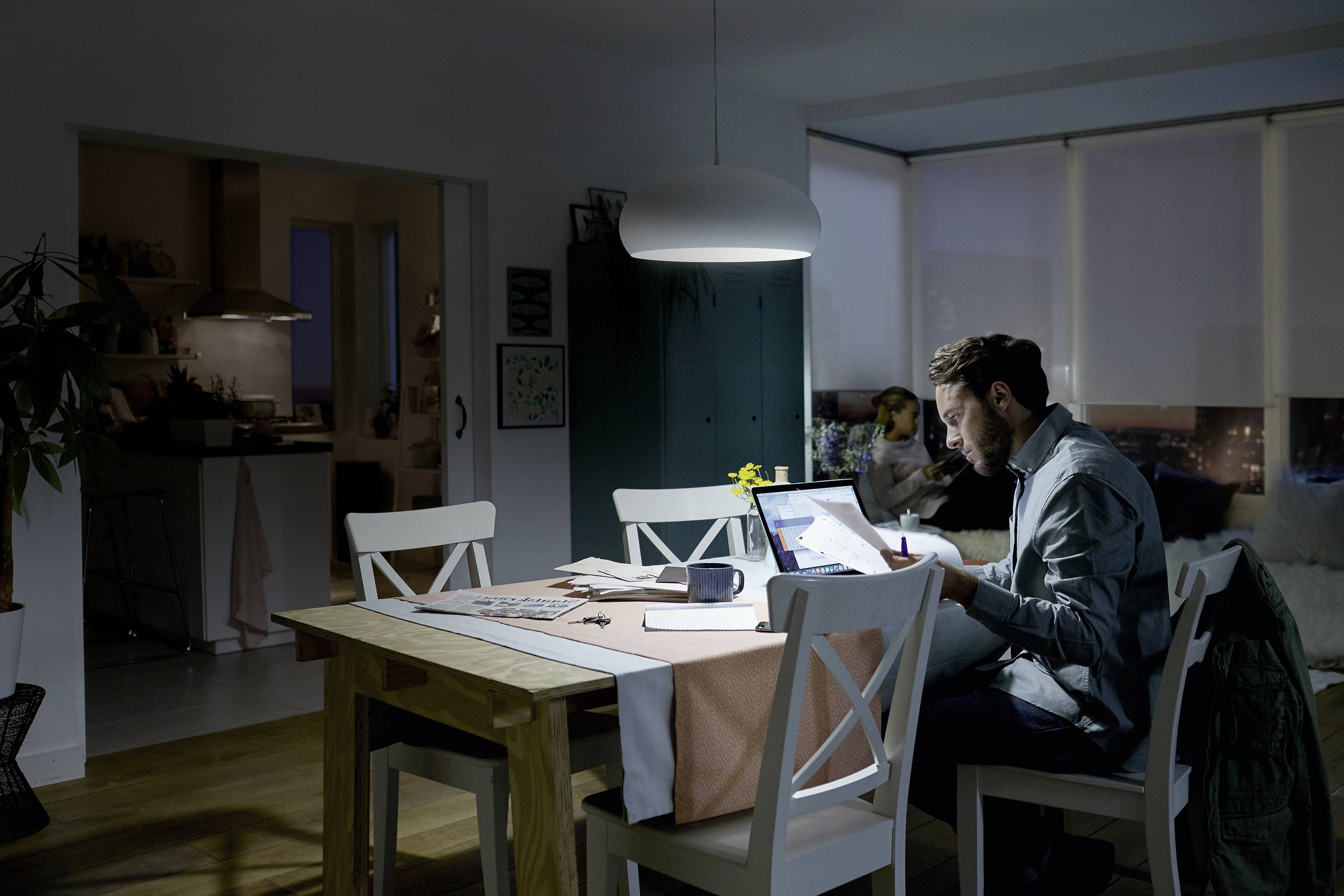 Ein Mann arbeitet abends mit einem Laptop am Esstisch, während eine Frau im Hintergrund auf dem Sofa sitzt. Die Beleuchtung ist gedämpft.
