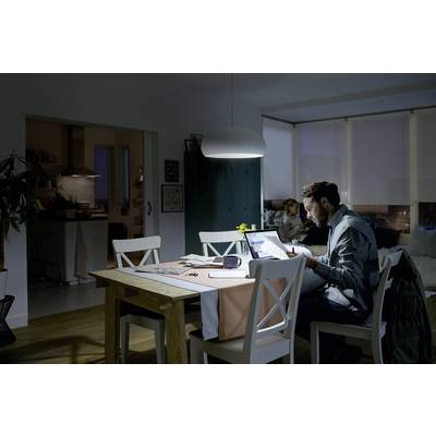 Ein Mann arbeitet abends mit einem Laptop am Esstisch, während eine Frau im Hintergrund auf dem Sofa sitzt. Die Beleuchtung ist gedämpft.