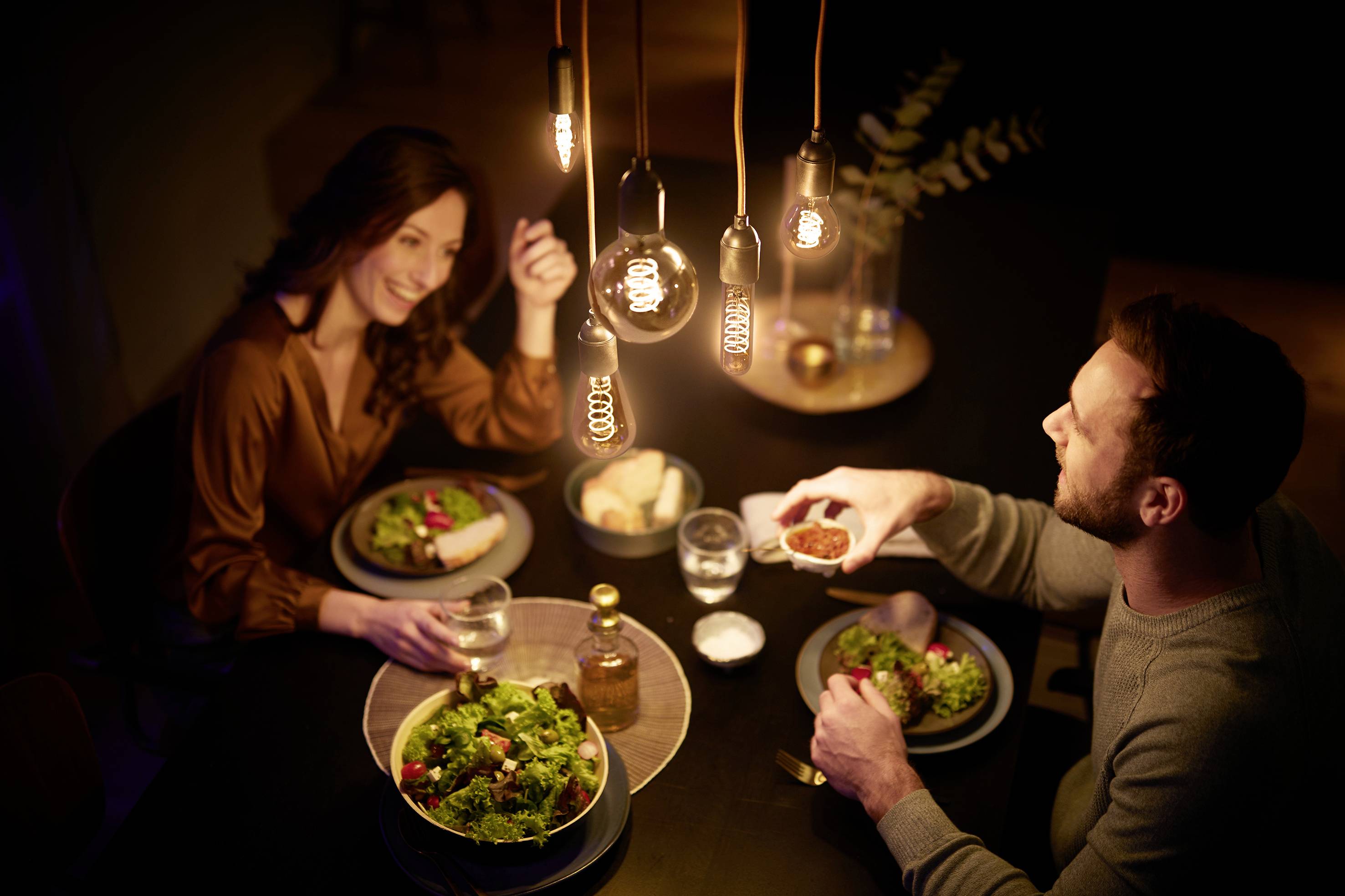 Ein Mann und eine Frau essen gemeinsam Abendessen in einem gemütlich beleuchteten Raum. Auf dem Tisch stehen Salate und Getränke.