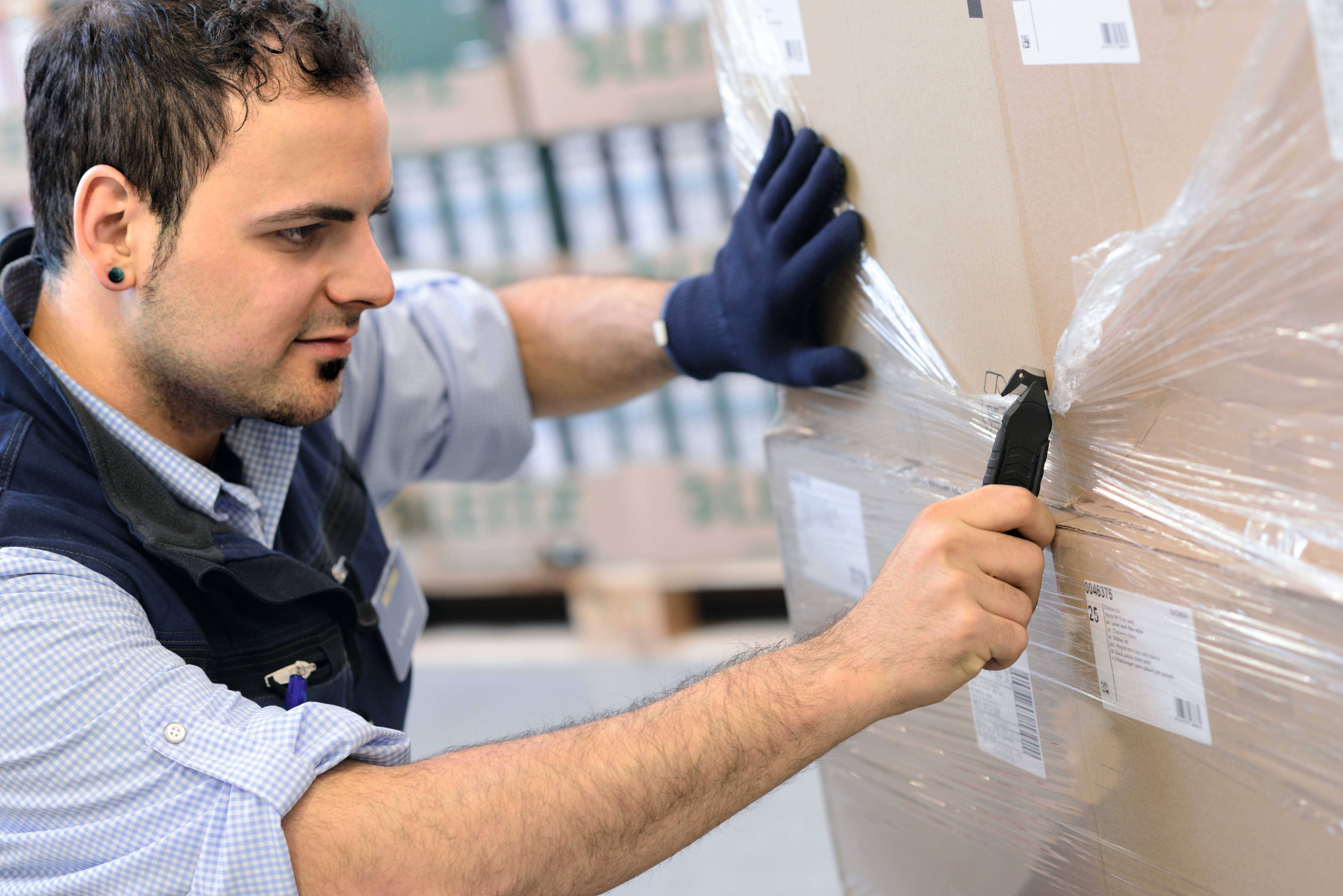 Ein Mann mit Handschuhen und einem Messer öffnet eine in Plastikfolie gewickelte Kiste in einem Lagerhaus.