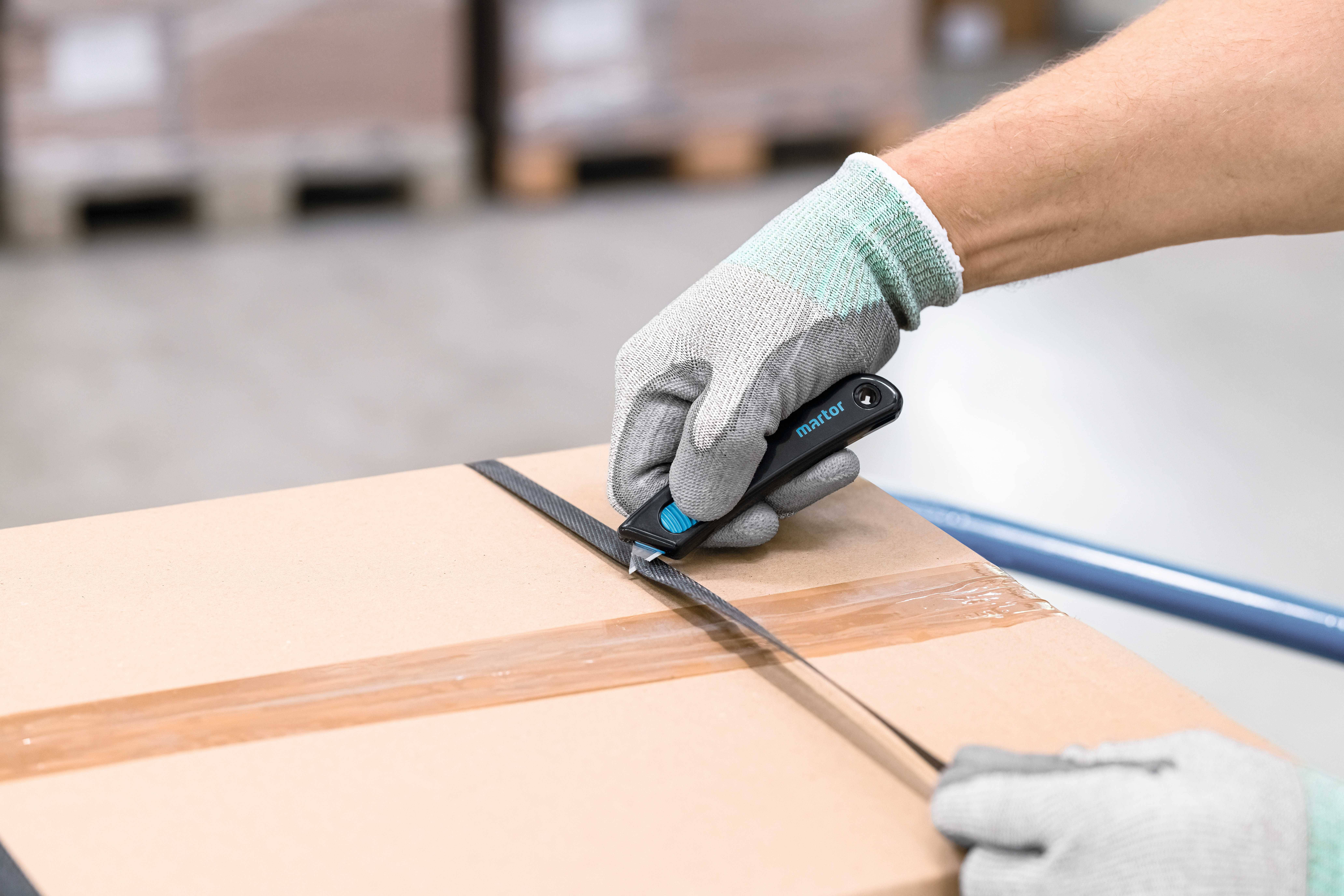 Eine Person mit grauen Handschuhen schneidet mit einem schwarzen Cutter ein Paketband auf einem Karton.