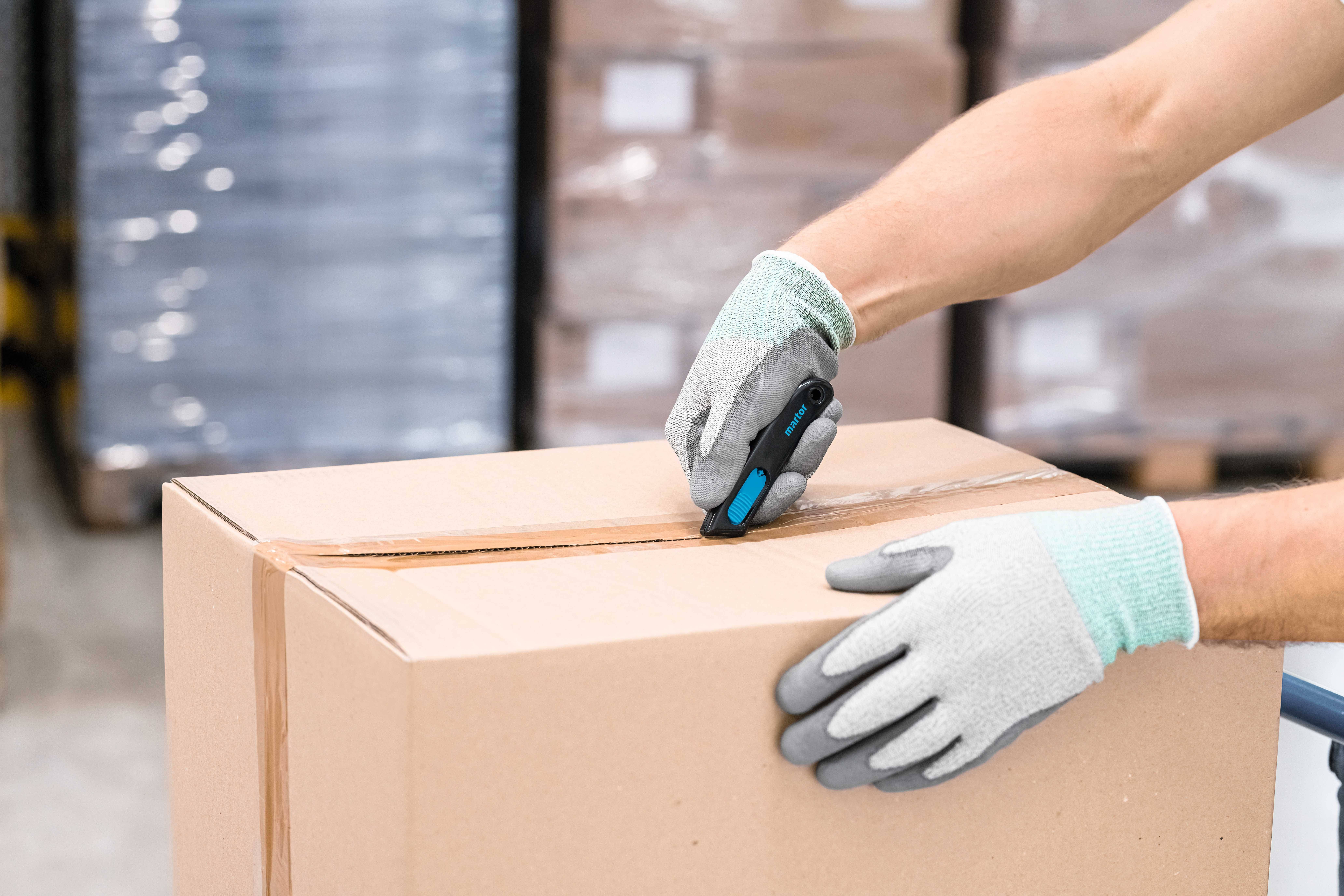 Person mit Handschuhen schneidet Karton mit Messer auf. Hintergrund zeigt gestapelte, plastikumwickelte Paletten.