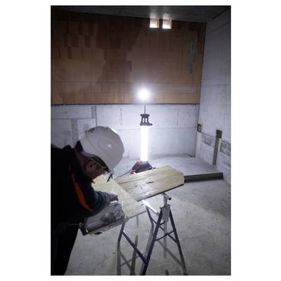 Arbeiter mit Schutzhelm, der ein Holzbrett auf Böcken in einem dunkel beleuchteten, unfertigen Raum zuschneidet, beleuchtet durch ein helles tragbares Arbeitslicht.