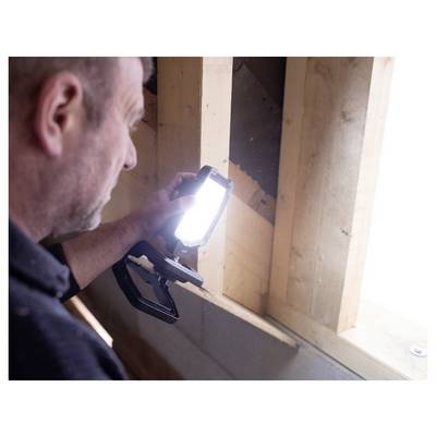 Ein Mann untersucht eine Wandöffnung mit einer tragbaren Arbeitsleuchte und überprüft die Holzkonstruktion im Inneren eines teilweise fertiggestellten Innenbereichs.