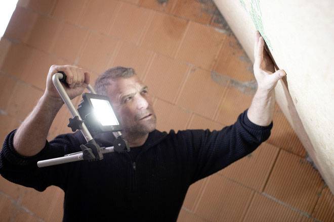 Ein Mann in schwarzem Pullover prüft bei hellem Licht eine Ziegelwand in einem Gebäude.