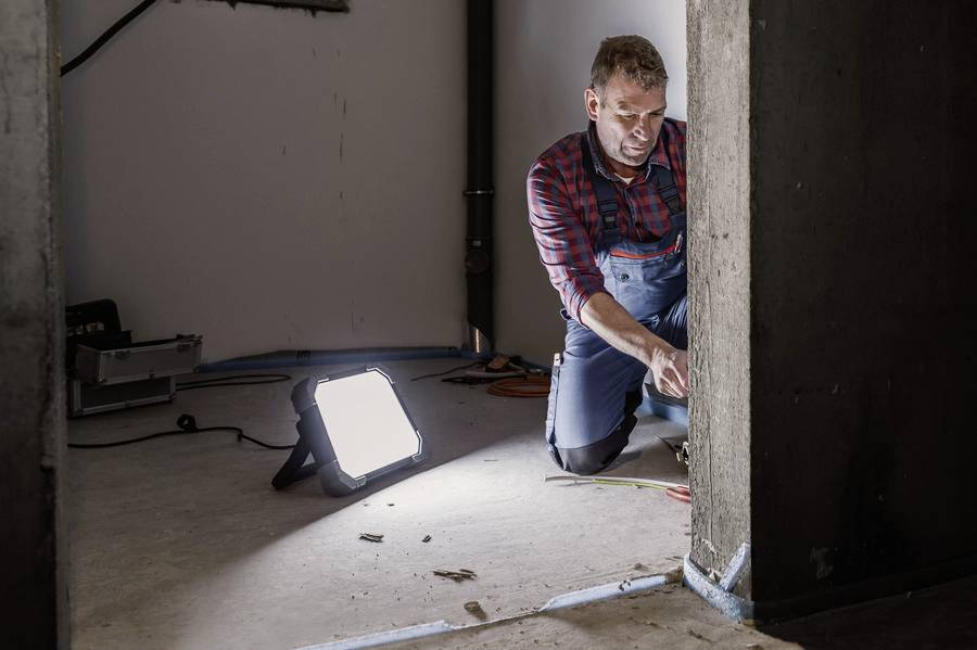 Ein Mann im Arbeitsanzug kniet und arbeitet an Kabeln in einem schlecht beleuchteten Raum. Eine helle Lampe beleuchtet den Bereich.