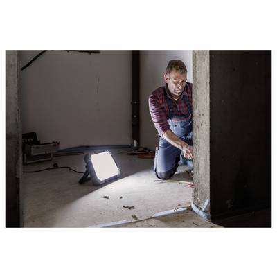 Ein Mann kniet neben einer Wand und nutzt Werkzeuge, um an Stromleitungen oder Reparaturen zu arbeiten, beleuchtet durch ein helles tragbares Arbeitslicht in einem unvollendeten Raum.