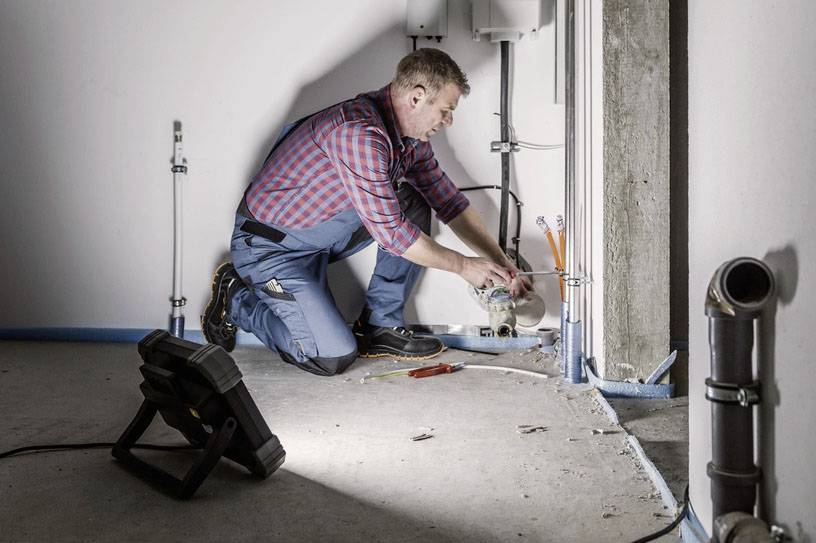 Ein Installateur kniet auf dem Boden und repariert Rohrleitungen in einem Raum. Werkzeug liegt neben ihm.
