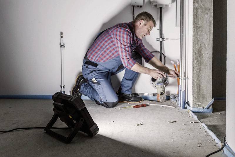 Ein Handwerker kniet in einem Raum und installiert Rohre an einer Wand. Im Vordergrund steht eine Lampe zur Beleuchtung.