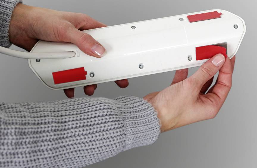 'Zwei Hände halten einen weißen Gegenstand mit roten Markierungen, der wie eine Verlängerungsschnur aussieht, gegen einen grauen Hintergrund.'