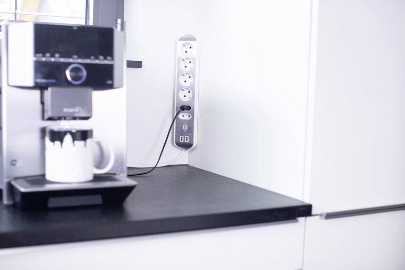 Eine Kaffeemaschine auf einer Küchenarbeitsplatte neben einer Steckdosenleiste mit digitalen Anzeige.