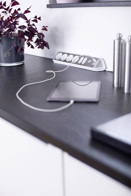 Tablet auf Tisch mit Ladekabel, verbunden mit einer Steckdosenleiste. Links eine Pflanze, rechts silberne Zylinderobjekte.