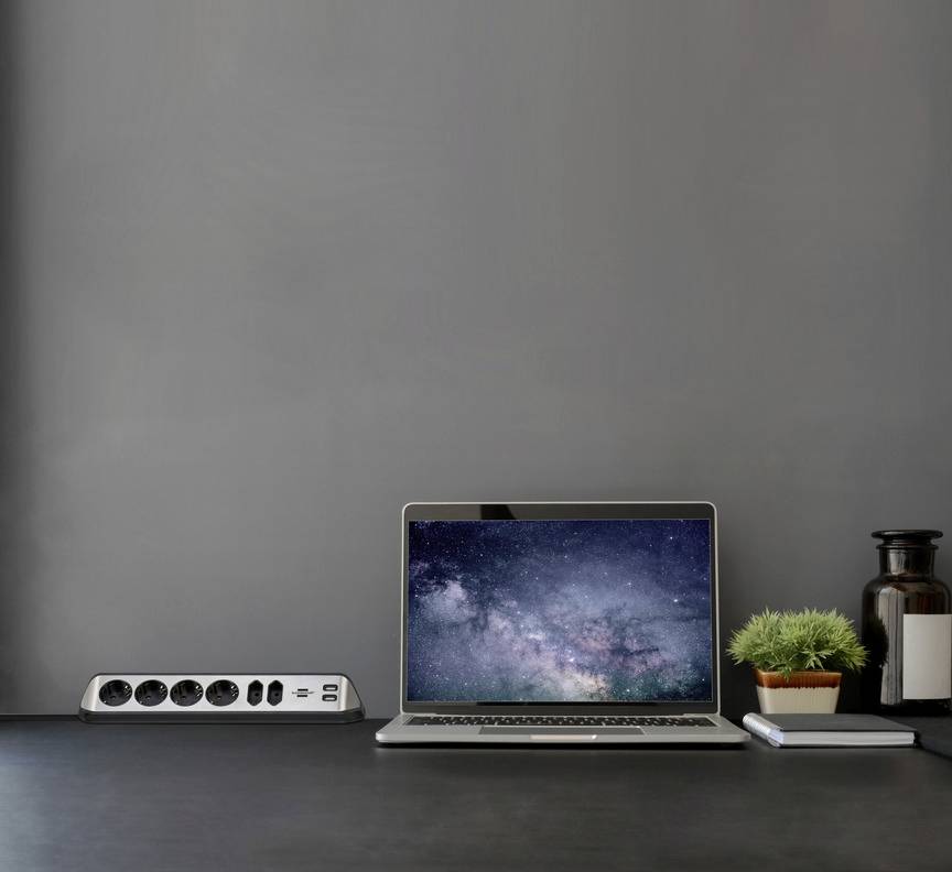 Ein Laptop auf einem Schreibtisch zeigt ein Bild der Milchstraße. Daneben stehen eine grüne Pflanze und eine Steckdosenleiste.