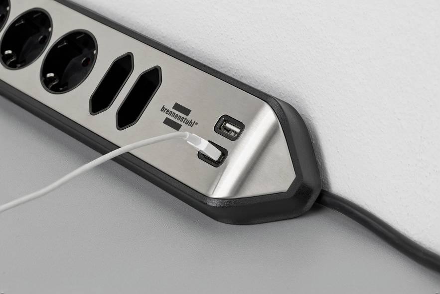 Steckdosenleiste mit zwei USB-Anschlüssen, ein weißes Kabel ist an einen der USB-Ports angeschlossen, auf grauer Oberfläche.