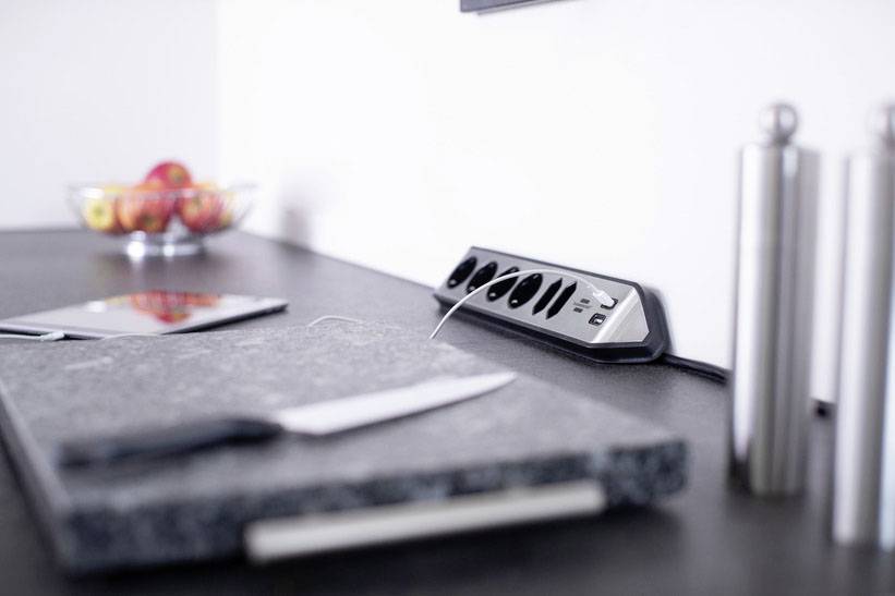 Eine moderne Küche mit einem Schneidebrett, Messer, Tablet auf der Arbeitsplatte und einer Steckdosenleiste an der Wand. Obstschale im Hintergrund.