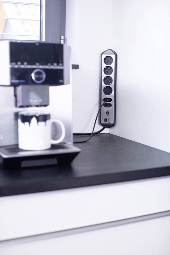Kaffeemaschine mit gefüllter Tasse auf schwarzer Arbeitsplatte; daneben Mehrfachsteckdose an der weißen Wand.