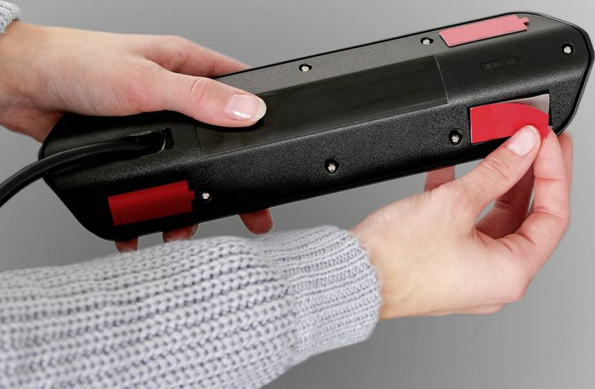 Eine Person hält ein schwarzes, rechteckiges Gerät mit roten Aufklebern auf der Rückseite, wahrscheinlich für eine technische Anwendung.