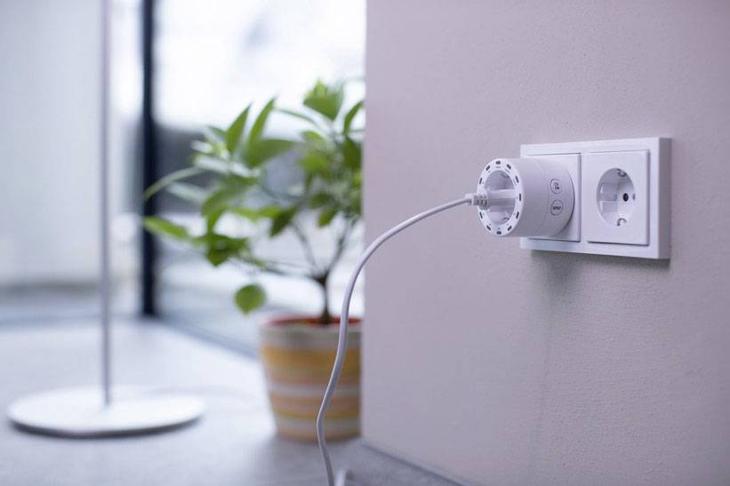 Ein weißes Gerät ist an eine Steckdose angeschlossen; im Hintergrund ist eine Topfpflanze sichtbar.