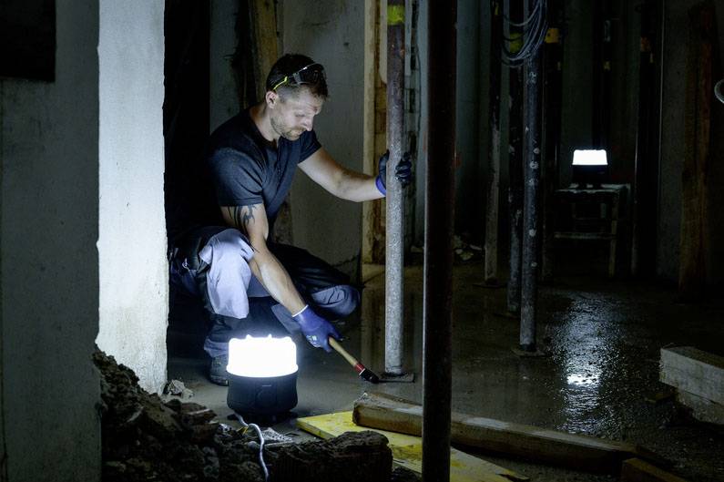 Ein Mann arbeitet mit einem Hammer in einem schwach beleuchteten Raum. Eine helle Lampe beleuchtet den Bereich, in dem er arbeitet.