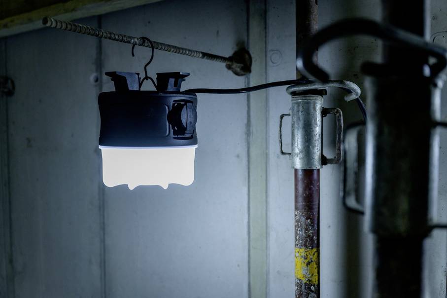 Eine helle LED-Lampe hängt an einem Haken in einem industriellen Raum, beleuchtet Rohre und eine Wand.