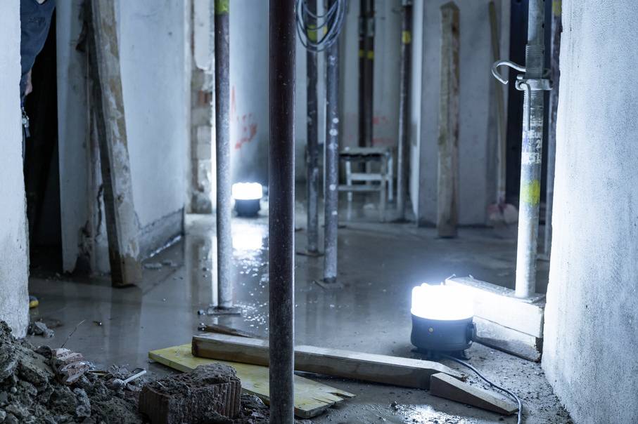 Baustelle mit nassen Böden und hellen Bauleuchten. Mehrere Stützen stabilisieren die Decke. Atmosphäre rau und industriell.