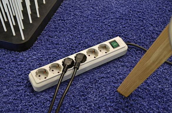 Ein verlängerungskabel auf blauem Teppich mit mehreren Steckdosen, von denen zwei mit schwarzen Kabeln belegt sind. Ein Holzbein eines Möbelstücks ist am Rand sichtbar.