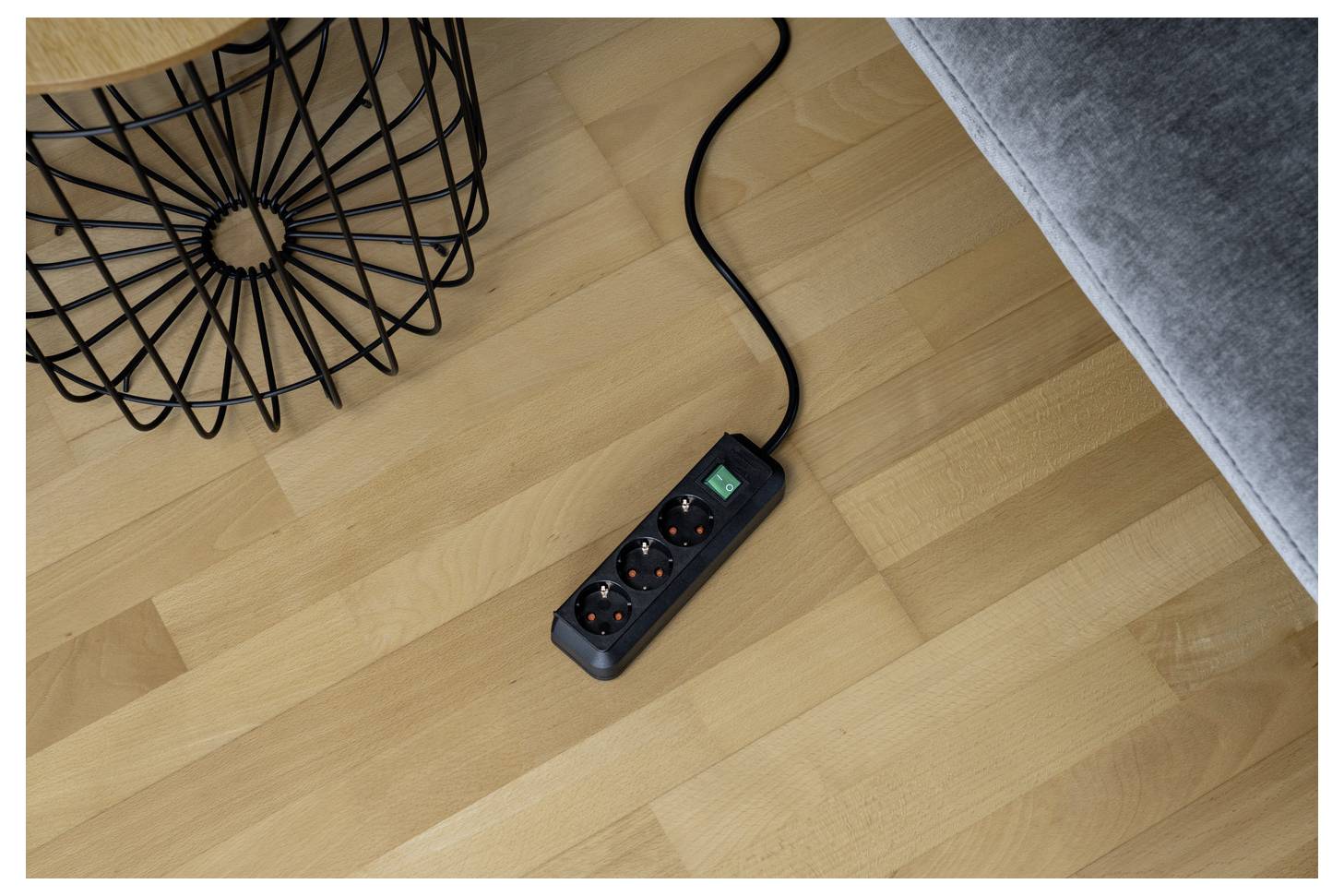 Schwarze Steckerleiste mit drei Steckdosen und einem beleuchteten Schalter auf einem Holzboden neben einem Sofa und einem Tischbein aus Drahtkorb.