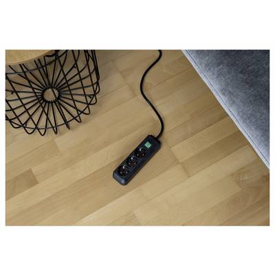 Schwarze Steckerleiste mit drei Steckdosen und einem beleuchteten Schalter auf einem Holzboden neben einem Sofa und einem Tischbein aus Drahtkorb.