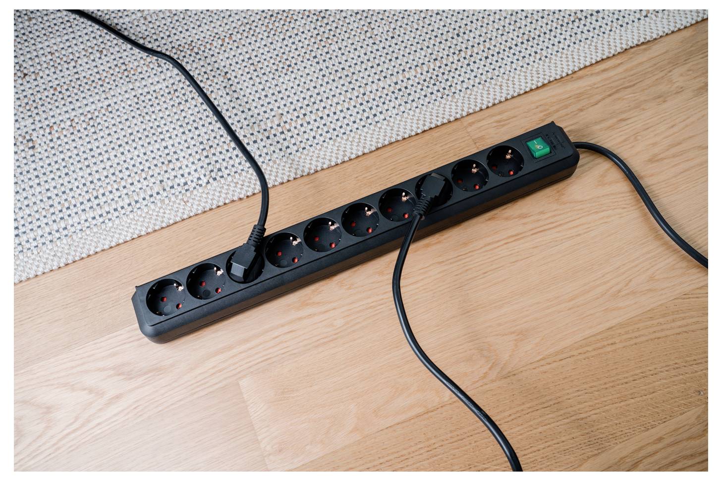 Schwarze Steckdosenleiste mit mehreren eingesteckten Kabeln auf einem Holzboden neben einer Wand.