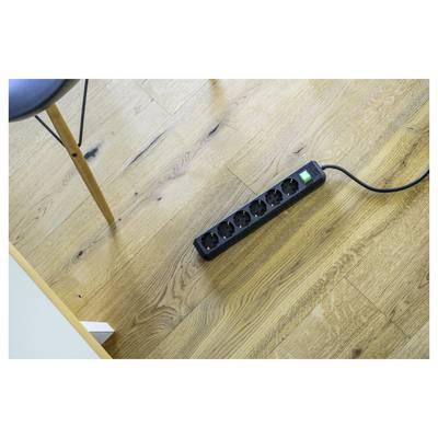 Eine schwarze Steckdosenleiste mit sechs Steckdosen und einem leuchtenden grünen Schalter liegt auf einem Holzboden neben einem Stuhlbein.