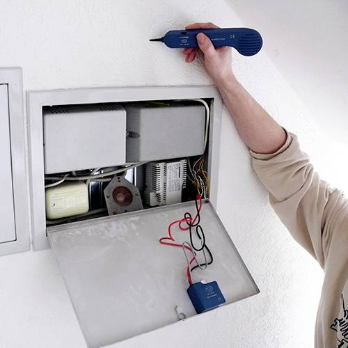 Person repariert eine elektronische Anlage in einer Wandöffnung mit Werkzeug. Gerät wird verkabelt, verschiedene Komponenten sichtbar.