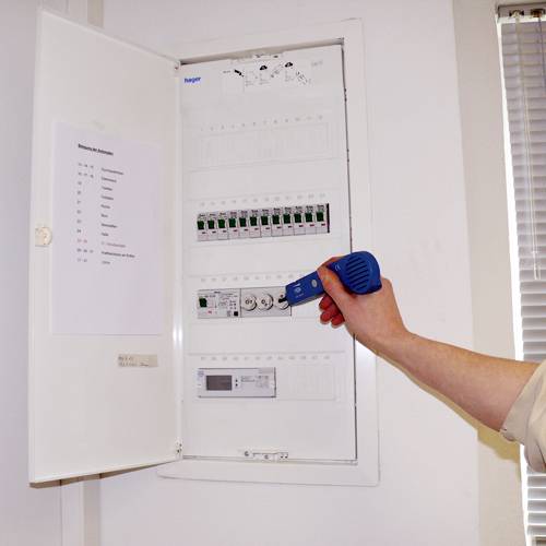 Eine Person überprüft mit einem elektronischen Gerät die Schaltkreise eines geöffneten Sicherungskastens im Innenbereich.