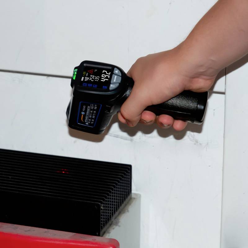 Eine Hand hält ein digitales Thermometer, das auf einen schwarzen Lüftungsschlitz zeigt. Das Display zeigt 27,6 Grad Celsius an.