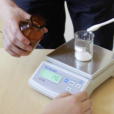 Ein Glas mit weißem Pulver wird auf einer elektronischen Waage gewogen. Eine Hand hält ein braunes Fläschchen. Gewichtsanzeige: 14,6 g.