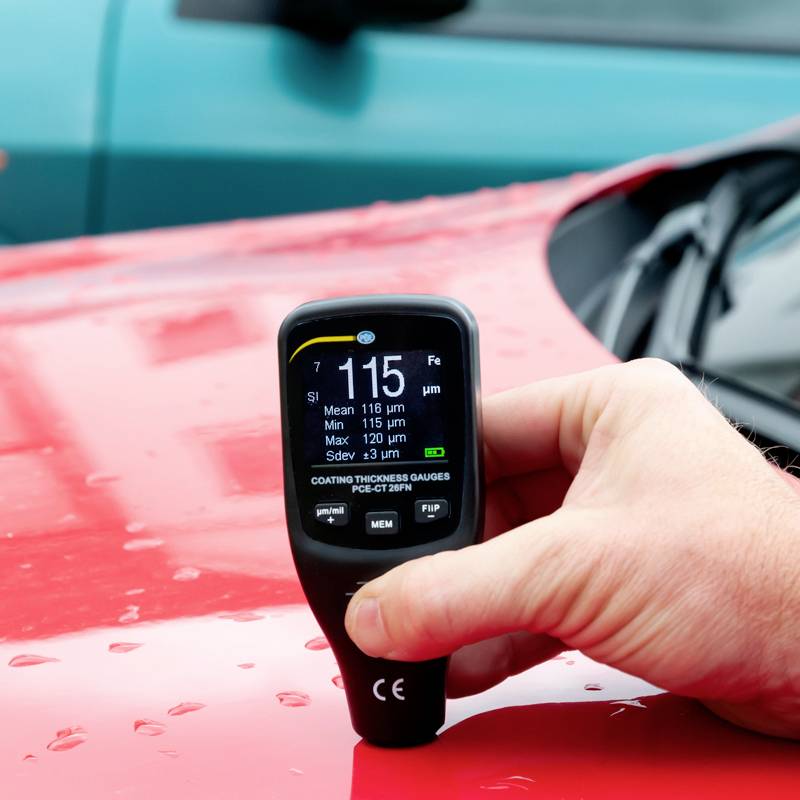 Eine Hand hält ein Schichtdickenmessgerät auf einer roten Fahrzeugoberfläche. Das Display zeigt Messwerte an. Im Hintergrund ein blaues Auto.