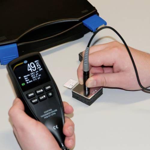 Zwei Hände bedienen ein Messgerät. Eine Hand hält das Gerät, das einen Wert anzeigt, die andere Hand führt eine Sonde an ein Testobjekt.