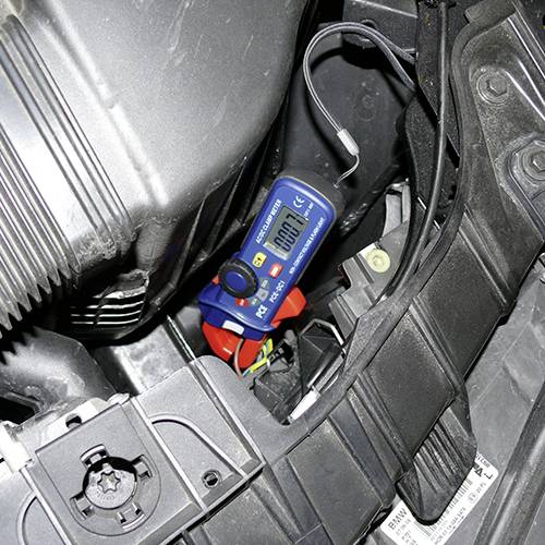 Ein Spannungsmessgerät zeigt 12,4 Volt an und ist an eine Autobatterie angeschlossen. Umgebung zeigt Teile eines Motorraums.