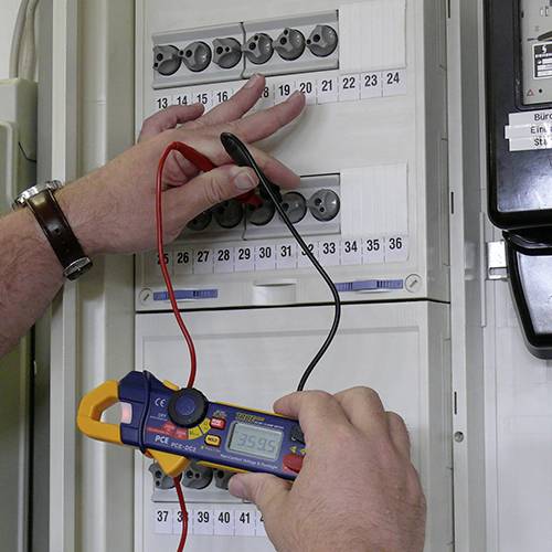 Eine Person misst mit einem Multimeter den Strom an einer elektrischen Schalttafel. Die Anzeige zeigt '39,95'.