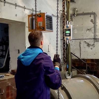 Eine Person in Arbeitskleidung bedient einen Kran in einer Werkshalle, hebt einen großen Metallzylinder mithilfe einer digitalen Waage.
