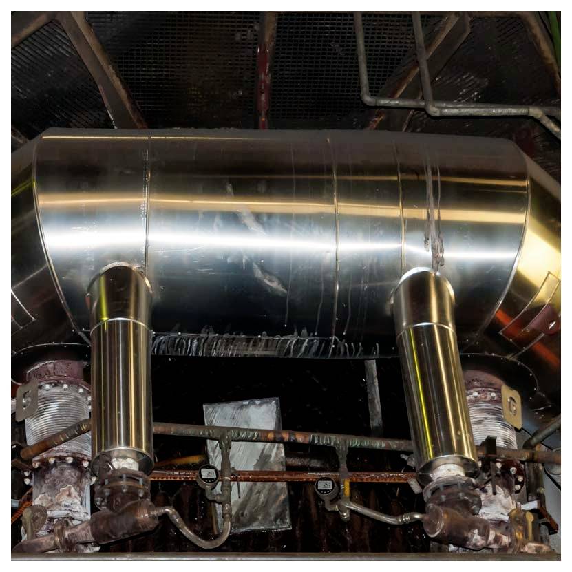 Glänzendes Metallausblaserohr und Rohre in einem industriellen Maschinenraum, mit zwei vertikalen Stützpfeilern und umgebender Maschinerie.