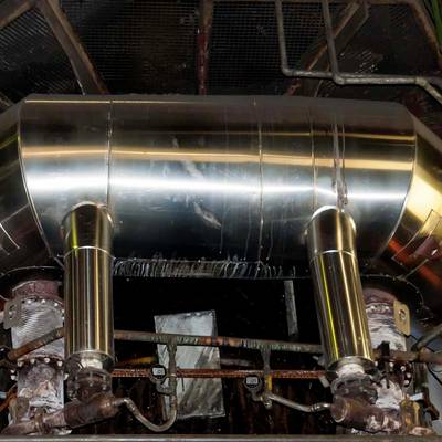 Ein großer, industrieller Metallzylinder mit zwei hervortretenden Rohren ist zentral montiert, umgeben von Rohrleitungen und Ventilen.