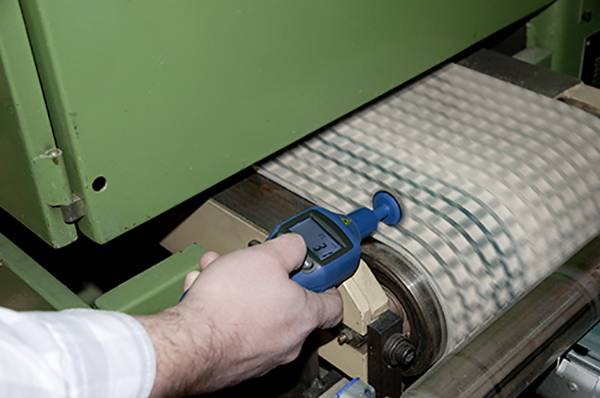 Eine Person misst mit einem blauen Messgerät die Dicke eines Materials auf einem Förderband. Hintergrund zeigt eine grüne Maschine.