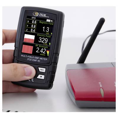 Hand hält ein Multi-Feld-EMF-Messgerät, das Magnetfeld-, Elektrofeld- und HF-Stärkemessungen zeigt, neben einem roten elektronischen Gerät mit einem Kabel.