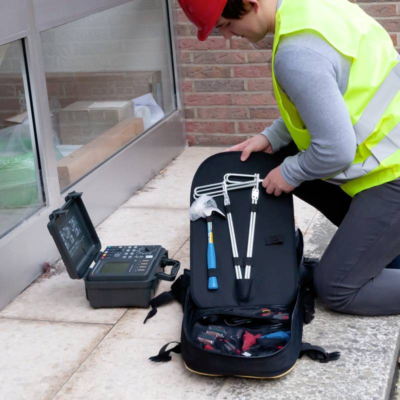 Ein Arbeiter in Warnweste und Helm bedient ein Messgerät vor einem Gebäude. Er hat Werkzeuge und ein offenes Gerät in der Nähe.